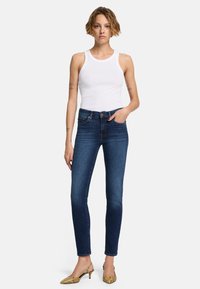 Dünne Jeans in dunkelblauem Denim mit einer glatten Textur. Kombiniert mit einem weißen Tanktop und goldenen Schlangenleder-High Heels. Enganliegendes Design.