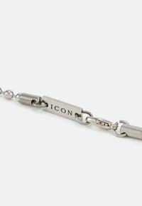 Colar de corrente de metal prateado com contas arredondadas, apresentando uma etiqueta retangular plana gravada com "ICON" e um fecho em uma das extremidades.