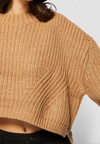 Gruby beżowy sweter z szerokim, prążkowanym dekoltem i luźnym krojem. Wzór teksturowany na całej powierzchni oraz krótki krój nad talią.