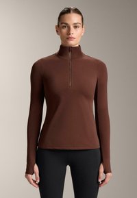 Top maro cu mânecă lungă, gât înalt și fermoar pe jumătate în față. Are o textură netedă și un design ajustat, asortat cu colanți negri.