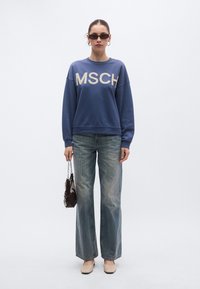 Μπλε φούτερ με το λογότυπο «MSCH», συνδυασμένο με ξεθωριασμένο τζιν παντελόνι φαρδιάς γραμμής. Αξεσουάρ περιλαμβάνουν μικρή καφέ τσάντα και σκούρα γυαλιά ηλίου.