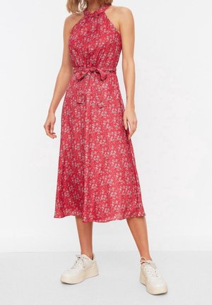 Robe rouge à col halter avec un motif floral, une taille cintrée à nouer et une longueur mi-mollet fluide. Associée à des baskets blanches.