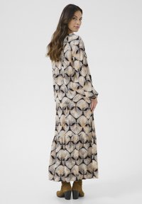 Maxikleid in Creme, Schwarz und Braun mit einem Blättermuster, mit langen Ärmeln, rundem Halsausschnitt und ausgestelltem Saum, kombiniert mit braunen kniehohen Stiefeln.