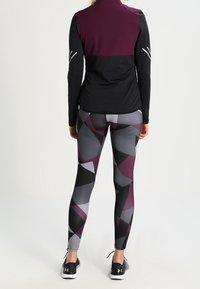 Una donna è in piedi con le spalle rivolte, indossando una maglietta a maniche lunghe di colore bordeaux e nero, leggings con motivo geometrico in nero, grigio e bordeaux, e scarpe da ginnastica nere.