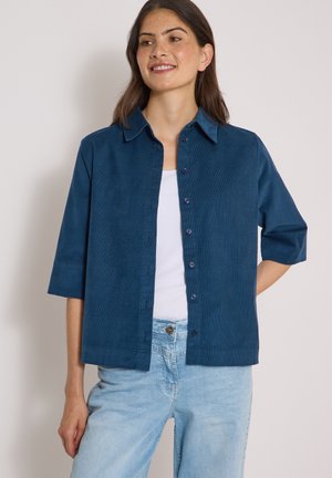Jonge vrouw draagt een blauw knoopjesoverhemd met ellebooglange mouwen over een wit topje en lichtblauwe spijkerbroek, staand met één hand achter haar rug.