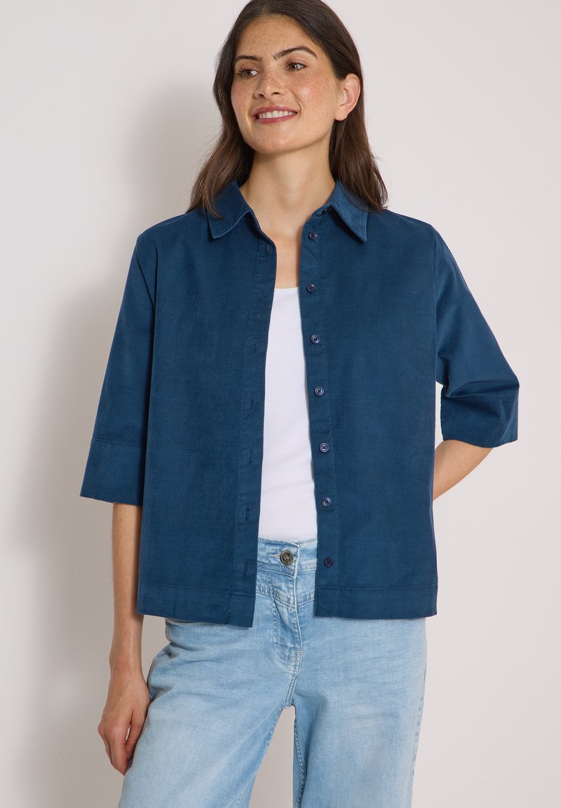 Jeune femme portant une chemise bleue boutonnée avec des manches jusqu'aux coudes sur un top blanc et un jean bleu clair, debout avec une main derrière le dos.
