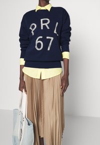 Navy sweatshirt with white print "PRL 67," layered over a light yellow collared shirt, paired with a pleated beige skirt and a blue tote bag.