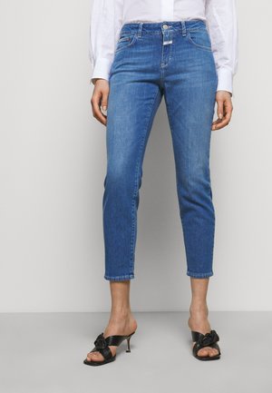 Kobieta ubrana w krótkie niebieskie obcisłe jeansy z wysokim stanem oraz czarne sandały na obcasie, stoi na jednolitym szarym tle.