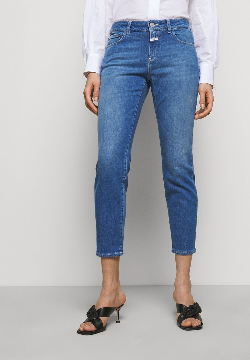 Femme portant un jean skinny bleu court taille haute et des sandales noires à talons, debout devant un fond gris uni.