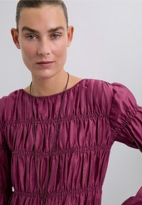 Fuchsia gerimpelde jurk met een ronde halslijn, lange mouwen en subtiele glans. Versterkt met een donkere ketting. Soepele textuur.