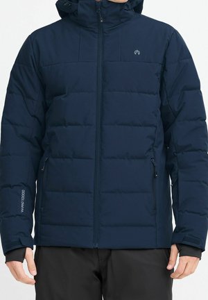 Granatowa, pikowana zimowa kurtka z przednim zamkiem błyskawicznym, kieszeniami na zamek i regulowanymi paskami na nadgarstkach, noszona przez osobę od szyi do bioder.