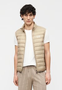 Colete acolchoado bege com gola alta, fecho de correr e logo no peito. Usado sobre uma camisa branca, combinado com calças bege com padrão.