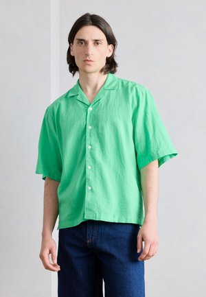 Νέο άτομο με σκούρα μαλλιά μέχρι τους ώμους, φορώντας ένα φωτεινό πράσινο πουκάμισο με κοντά μανίκια και σκουρόχρωμο μπλε τζιν, ουδέτερο φόντο.