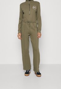 Sweatshirt zippé vert olive avec un col haut et un logo ; associé à un jogging assorti avec une taille élastique et des jambes évasées.