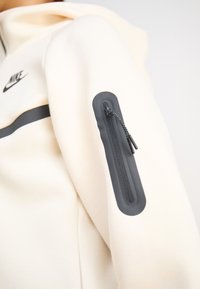 Hoodie beige claro com um bolso de zíper preto na manga, apresentando uma faixa preta ao longo do peito e um logo da Nike em preto.