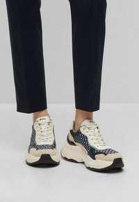 Sporty sneakers feature a mix of navy blue, cream, and gray materials with mesh detailing, laces, and a chunky sole. Branding visible on tongue.