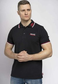 Fekete póló, piros és fehér kiegészítőkkel, gallérral és hímzett logóval. A modellen világoskék farmernadrág van.