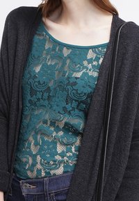 Teal spetsbodysuit lagerad under en mörkgrå kofta med blommönster och rundad halsringning. Mörka denimjeans fulländar looken.