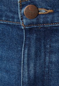 Tela de denim con un primer plano de un botón de cobre y costuras doradas en contraste; presenta una costura vertical y un color azul desvanecido.