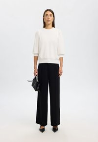 Weißer Pullover mit Puffärmeln, schwarze weite Hosen und spitze schwarze Pumps. Hält eine kleine schwarze Handtasche.