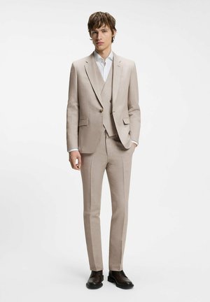 Joven vestido con un traje beige de tres piezas, con camisa blanca y zapatos de vestir negros, de pie con una mano en el bolsillo.