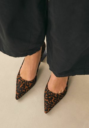 Pointed-toe heels in black and brown leopard print fabric, featuring a low-cut vamp and contrasting solid heel. Textured surface with a soft finish.