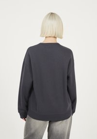 Persona con capelli biondo platino corti, indossa una felpa a maniche lunghe grigio scuro e pantaloni grigio chiaro, vista da dietro.