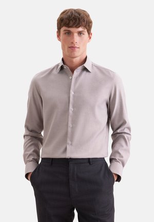 Jeune homme portant une chemise à manches longues gris clair boutonnée avec un pantalon sombre, debout les mains dans les poches devant un fond blanc.