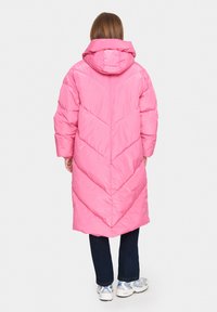 Hosszú rózsaszín puffertakó, cikkcakk mintával, kapucnival és túlméretezett ujjal, sötét farmernadrággal és szürke sportcipővel párosítva.