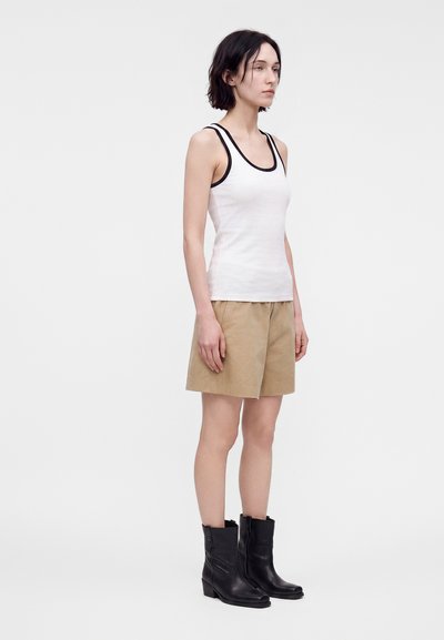 Femme portant un débardeur blanc avec un bord noir, un short beige jusqu'aux genoux, et des bottes noires mi-mollet, debout devant un fond clair uni.