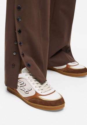 Valged ja pruunid spordijalatsid, millel on tekstuuriga kangast pealisosa, beeži tooni aktsendid ja kummist tallaosa, mille on kombineeritud vabalootud pruunide pükstega, millel on külgklambrid.