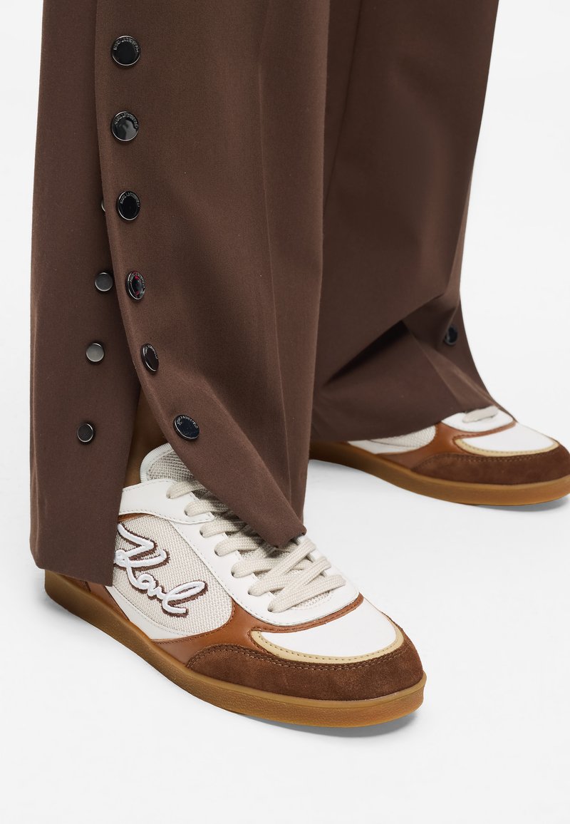 Fehér és barna sportcipők texturált szövet felsőrésszel, bézs részletekkel és gumitalppal, laza barna nadrággal párosítva, amely oldalsó gombokkal rendelkezik.