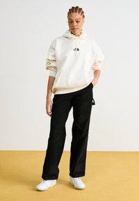 Felpa crema con coulisse e tasca frontale, abbinata a pantaloni larghi neri e sneakers bianche, il tutto su uno sfondo giallo.
