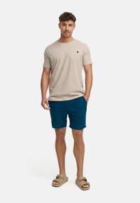 Beige T-Shirt mit kurzen Ärmeln und Tasche, kombiniert mit blauen Shorts und beigen Sandalen. Lässige, entspannte Passform und weiche Textur.