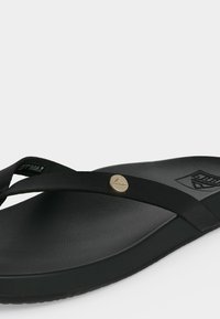 Μαύρο σανδάλι flip-flop με υφή στη σόλα, λεπτά λουράκια που διασταυρώνονται κοντά στα δάχτυλα και μικρό στρογγυλό μεταλλικό έμβλημα στο πάνω λουράκι.