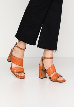 Frau trägt schwarze, kurze Hosen und orangefarbene High-Heel-Sandalen mit Knöchelriemen und strukturiertem Muster auf einem weißen Boden.