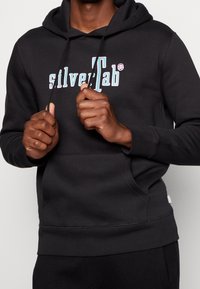 Svart huvtröja med framficka, snörning och tryckt logotyp i blått och rosa. Mjuk tyg med en avslappnad passform.