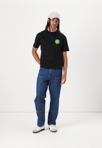 Black short-sleeve t-shirt with a circular green graphic and yellow design on the chest, paired with loose blue denim jeans and white sneakers.