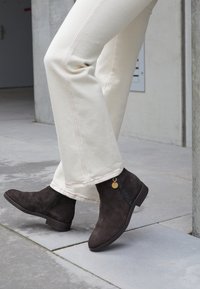 Stivali alla caviglia in suede marrone con zip laterale e un accento dorato decorativo. Abbinati a pantaloni color crema con cuciture a contrasto.