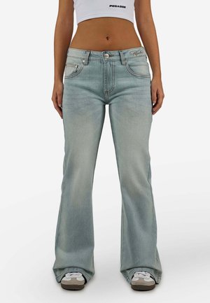 Vrouw draagt lichtblauwe uitlopende spijkerbroek, wit sportief crop top en witte sneakers, staand tegen een effen witte achtergrond.