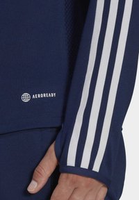 Marineblaue Sportbekleidung mit weißem dreisträngigen Design an den Ärmeln und Aeroready-Logo. Texturiertes Material, enganliegende Bündchen und Nahaufnahme einer Hand.