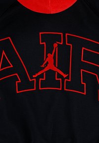 Tissu noir avec de grandes lettres bordées de rouge formant AIR et une silhouette rouge d’un joueur de basket sautant entre les lettres.