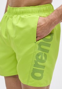 Pantalones cortos de baño verde lima con una cinturilla elástica, bolsillo lateral y un detalle de logo de malla negra a lo largo del lado izquierdo. Textura suave.