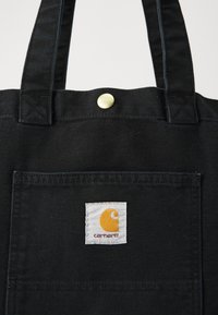 Borsone in canvas nero con due manici, dotato di una tasca frontale e una targhetta bianca con un logo giallo. Dettaglio della chiusura con snap in ottone.