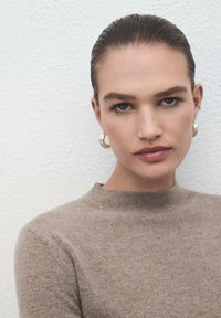 Suéter marrón de punto con un cuello alto, que presenta una textura sutil y acentos metálicos. La modelo tiene el cabello liso y un maquillaje neutro.