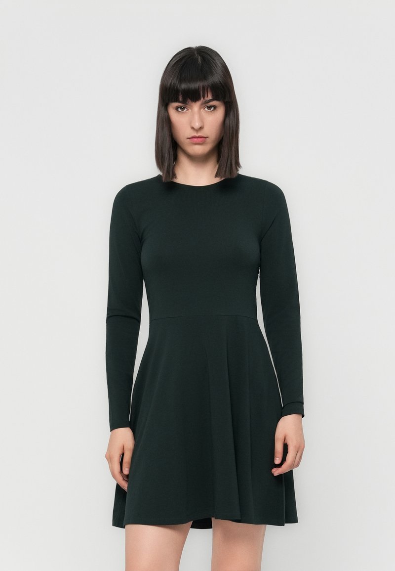 Robe longue verte à manches longues avec un corsage ajusté et une jupe évasée. Fabriquée en tissu doux avec une texture lisse et un col rond.