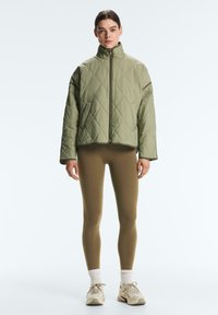 Kvinde iført grøn quiltet jakke, olivengrønne leggings, hvide sokker og beige sneakers, stående mod en ensfarvet lys baggrund.