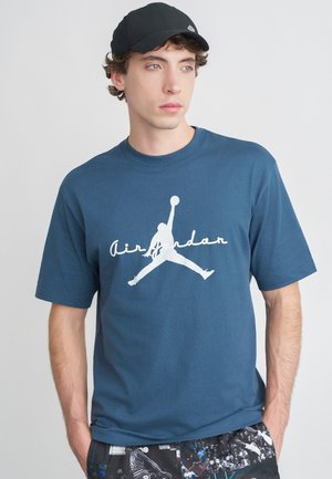 Μπλε σκούρο βαμβακερό μπλουζάκι με λευκό λογότυπο "Air Jordan" που απεικονίζει τη σιλουέτα ενός άλτη. Χαλαρή γραμμή, στρογγυλή λαιμόκοψη.