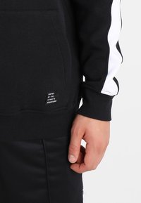 Svart hoodie med vita sidostreck, med en känguruficka och ribbade muddarna. Varumärkesetikett synlig på nedre vänstra sidan.