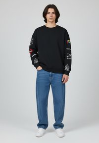Felpa nera con grafiche sulle maniche, abbinata a jeans in denim azzurro chiaro e sneaker bianche. Silhouette a vestibilità rilassata.